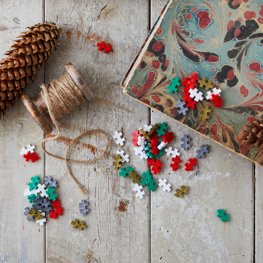 Red, green, white and gold Plus-Plus pieces scattered on wood with pinecones and twine